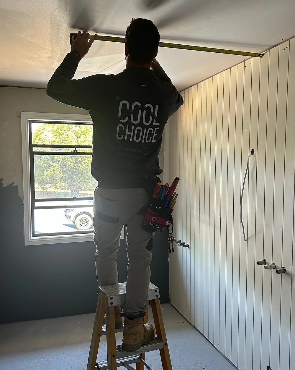 A Cool Choice electrician in a branded shirt standing on a step ladder measuring the ceiling of a new room