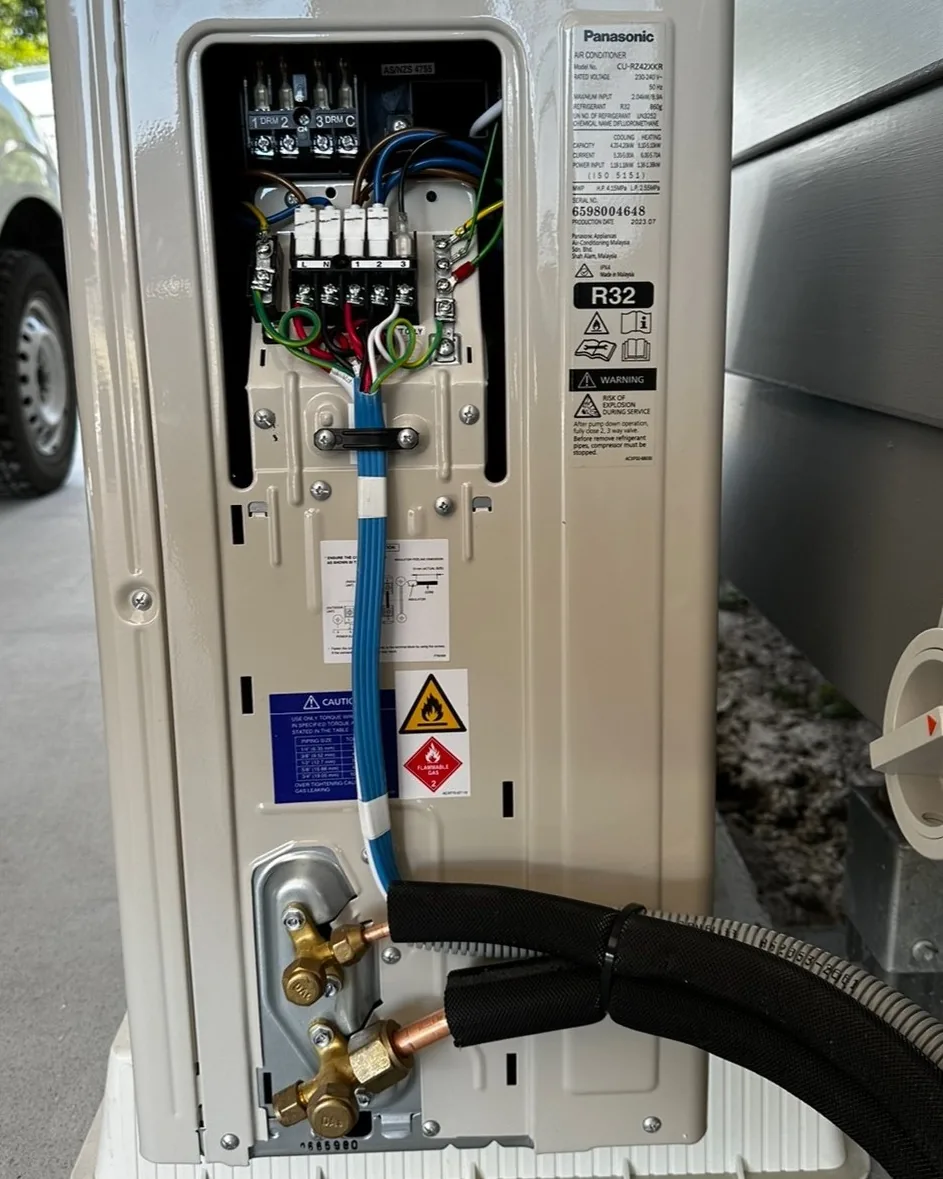 A Panasonic R32 outdoor unit with its terminal cover off showing neat wiring and pipework, a Cool Choice ute behind