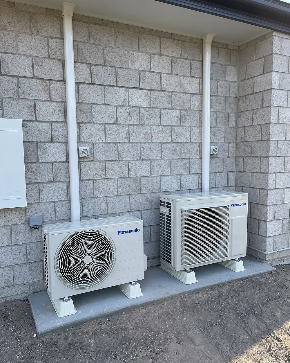 Two Panasonic split system outdoor units on brackets against a block wall, with white pipe covers running up the wall